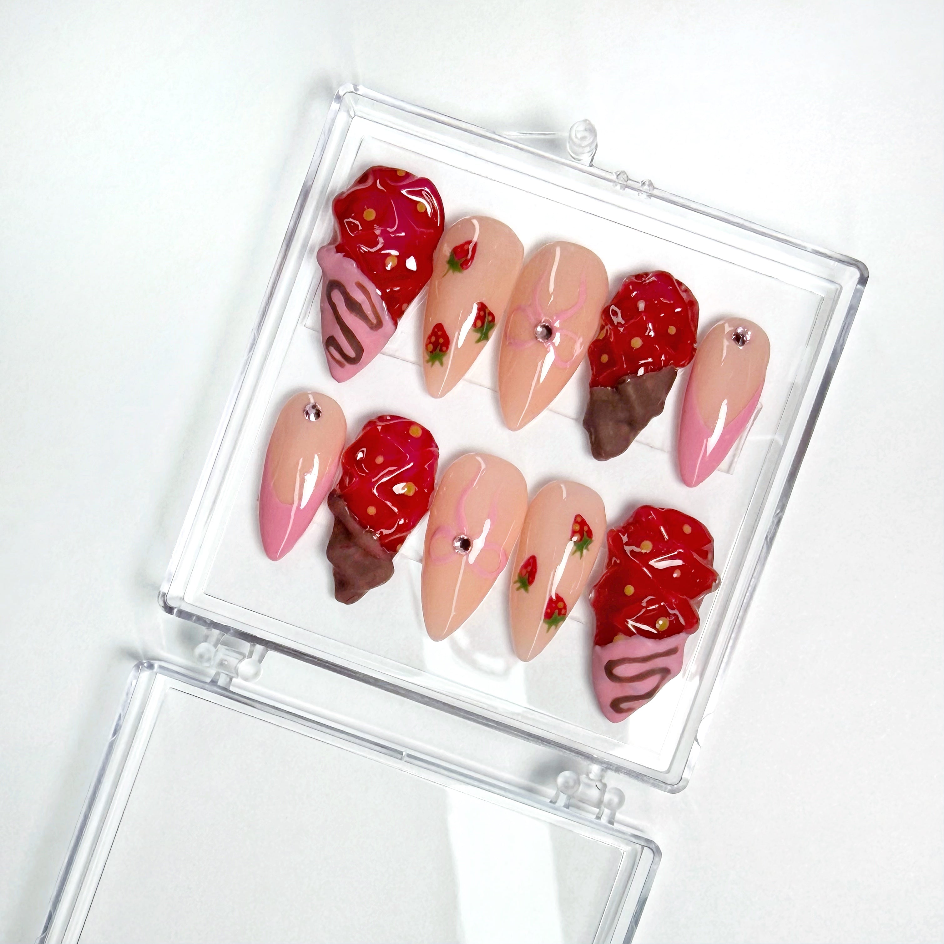 Set of decorative nail art with strawberry and chocolate drizzle designs in a clear case on a white background