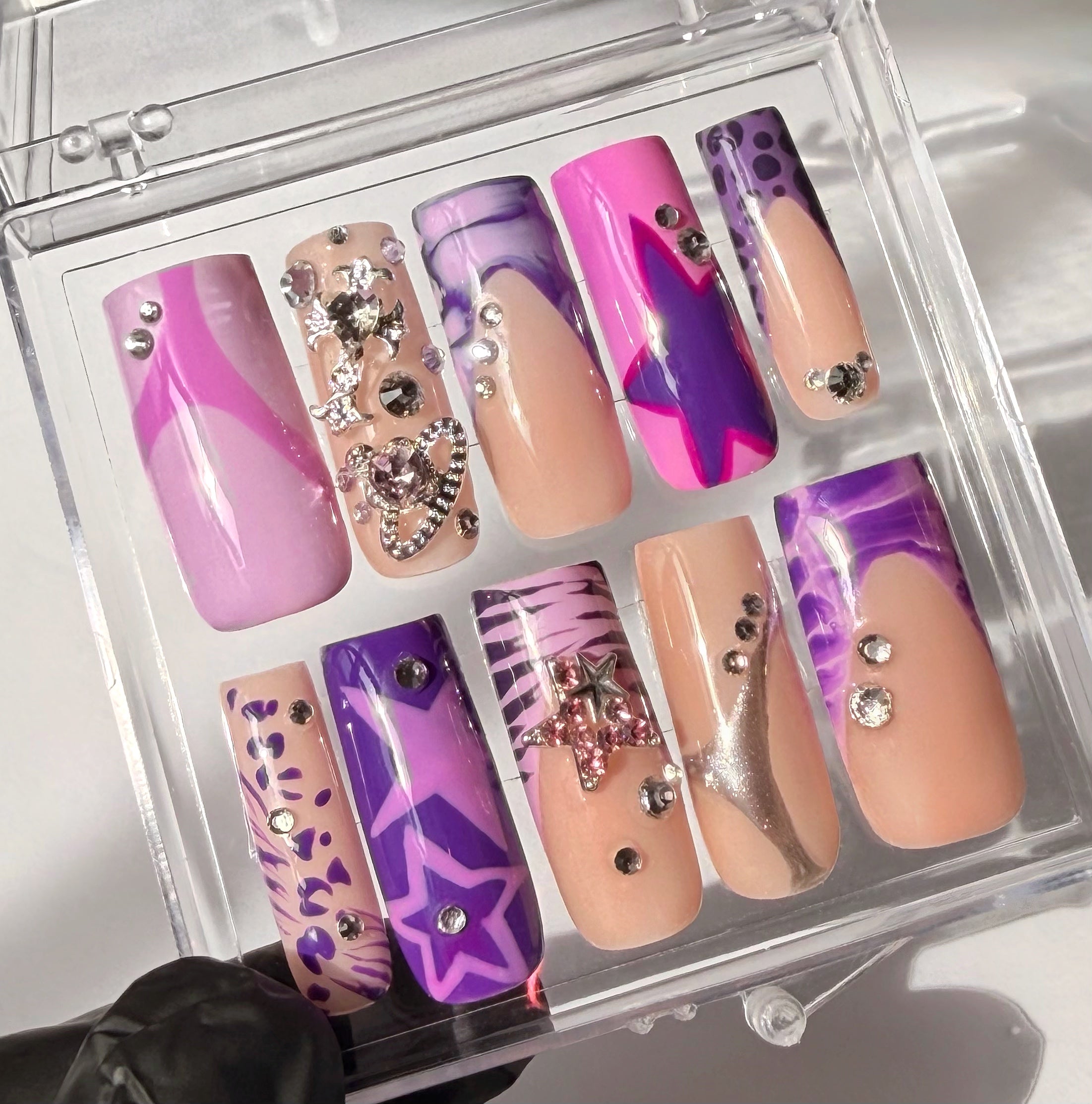 Set of decorative false nails with pink, purple, and beige designs in a clear display case.