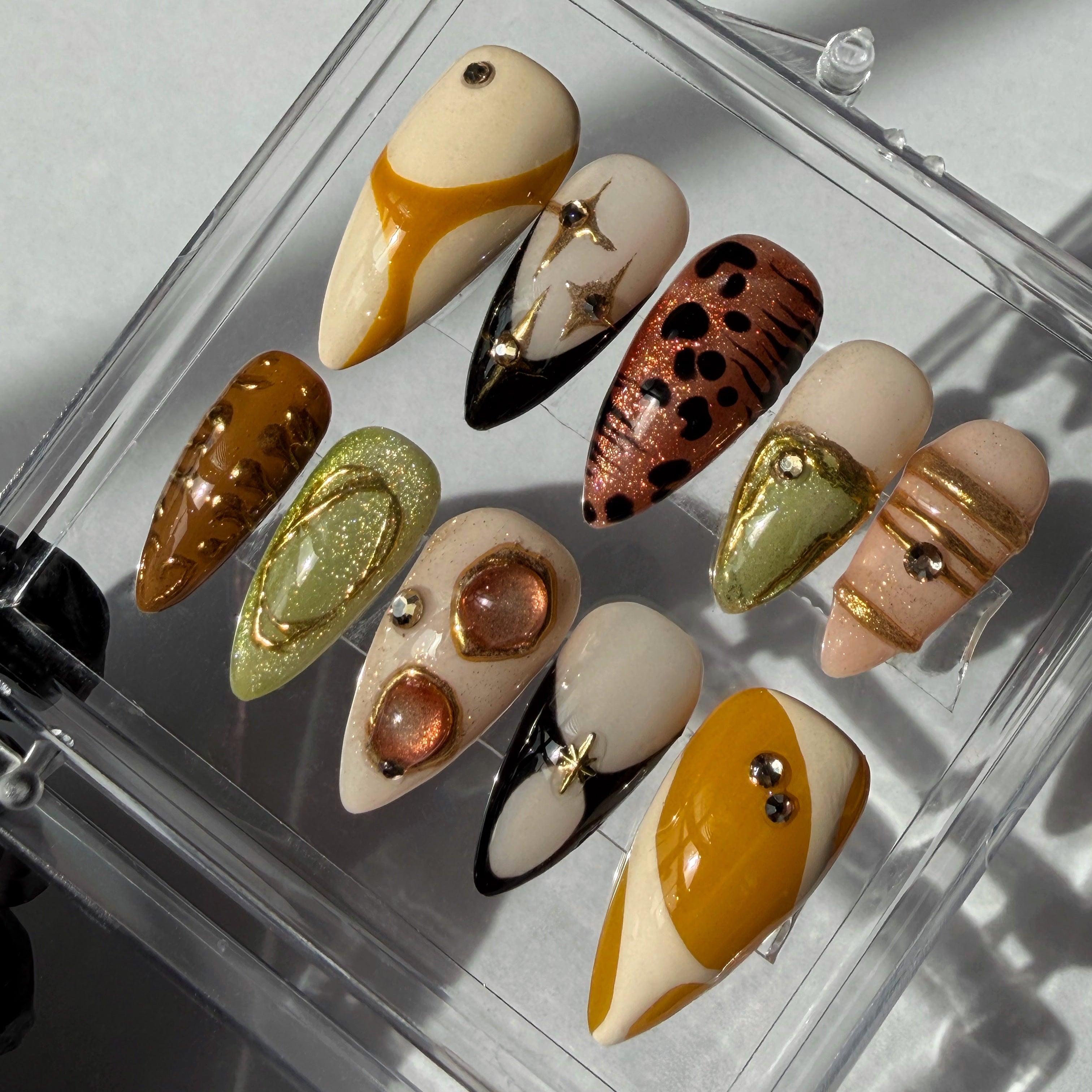 Set of decorative nail art tips with yellow tones, gold accents, black French tips, in a clear display case on a light background