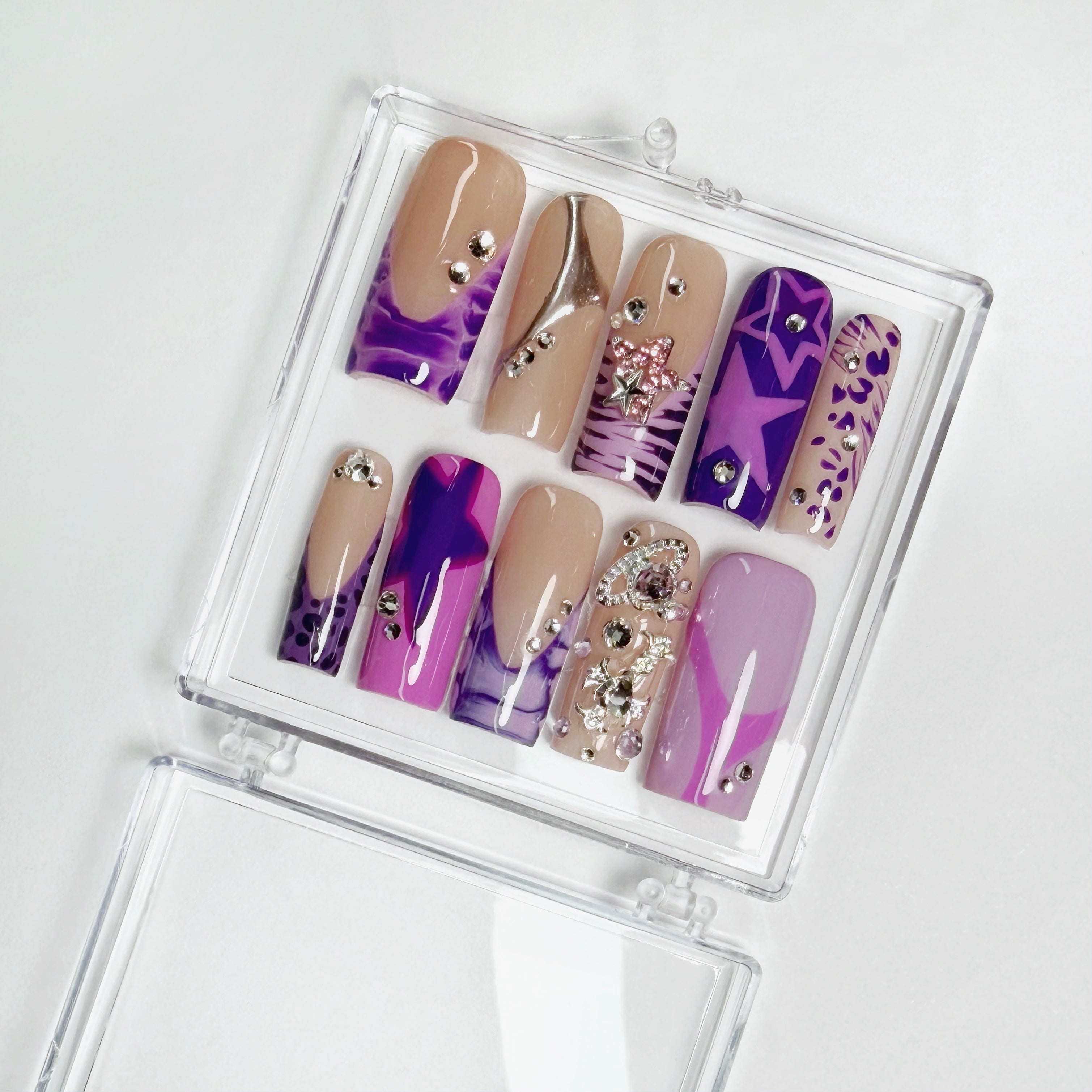 Set of purple and beige nail designs with rhinestones in a clear case on a white background