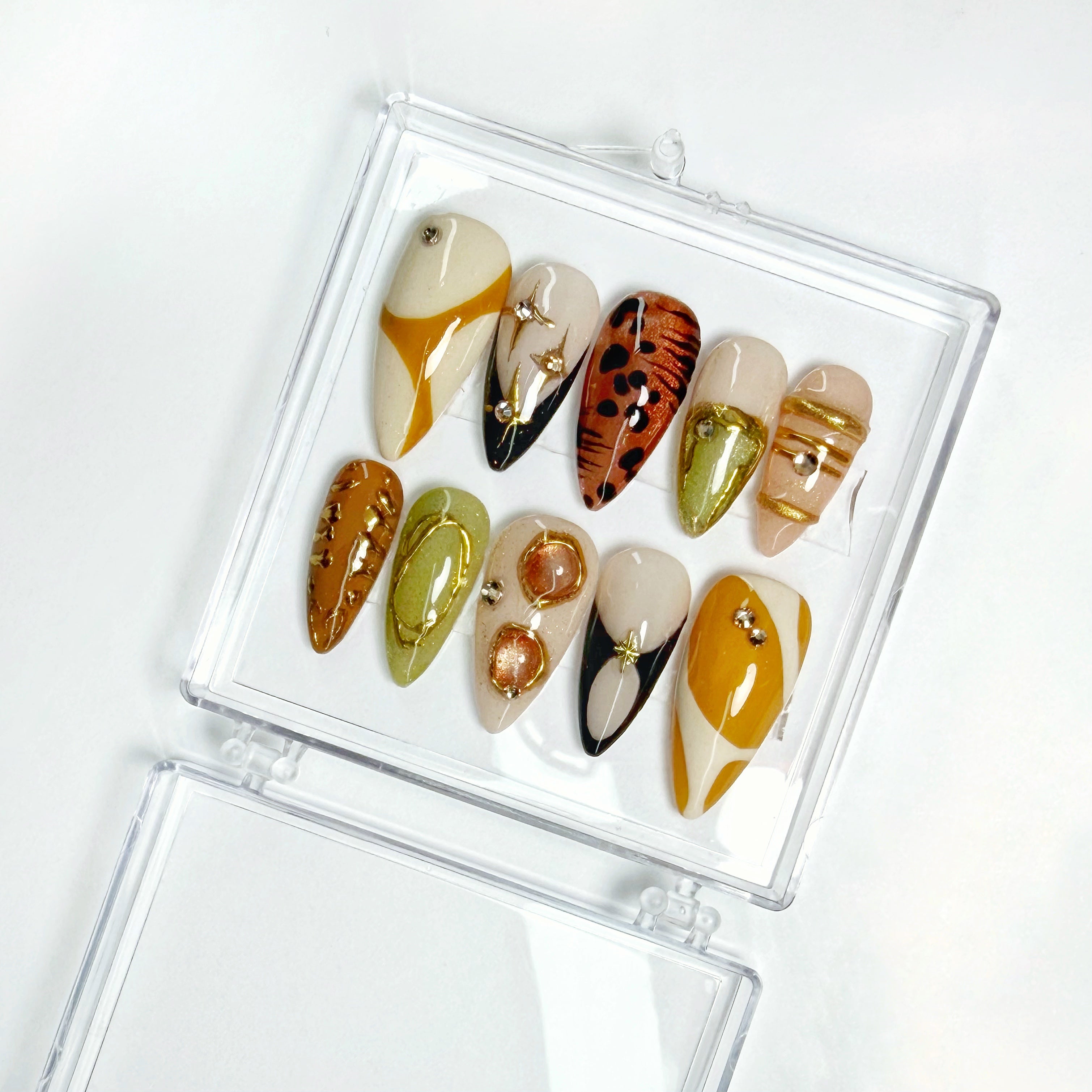 Brown, gold, and nude abstract press-on nail set with rhinestone accents in a clear display case.