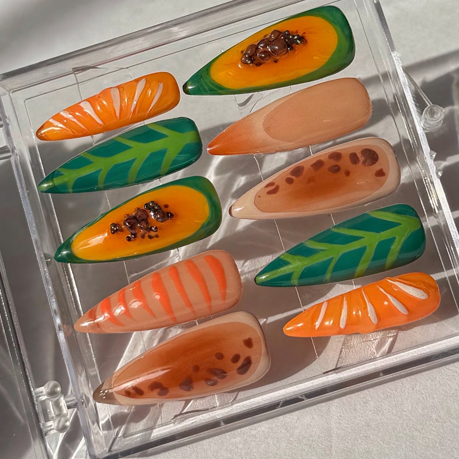 Set of papaya green, orange, and brown fruit-themed false nails in a clear display case.