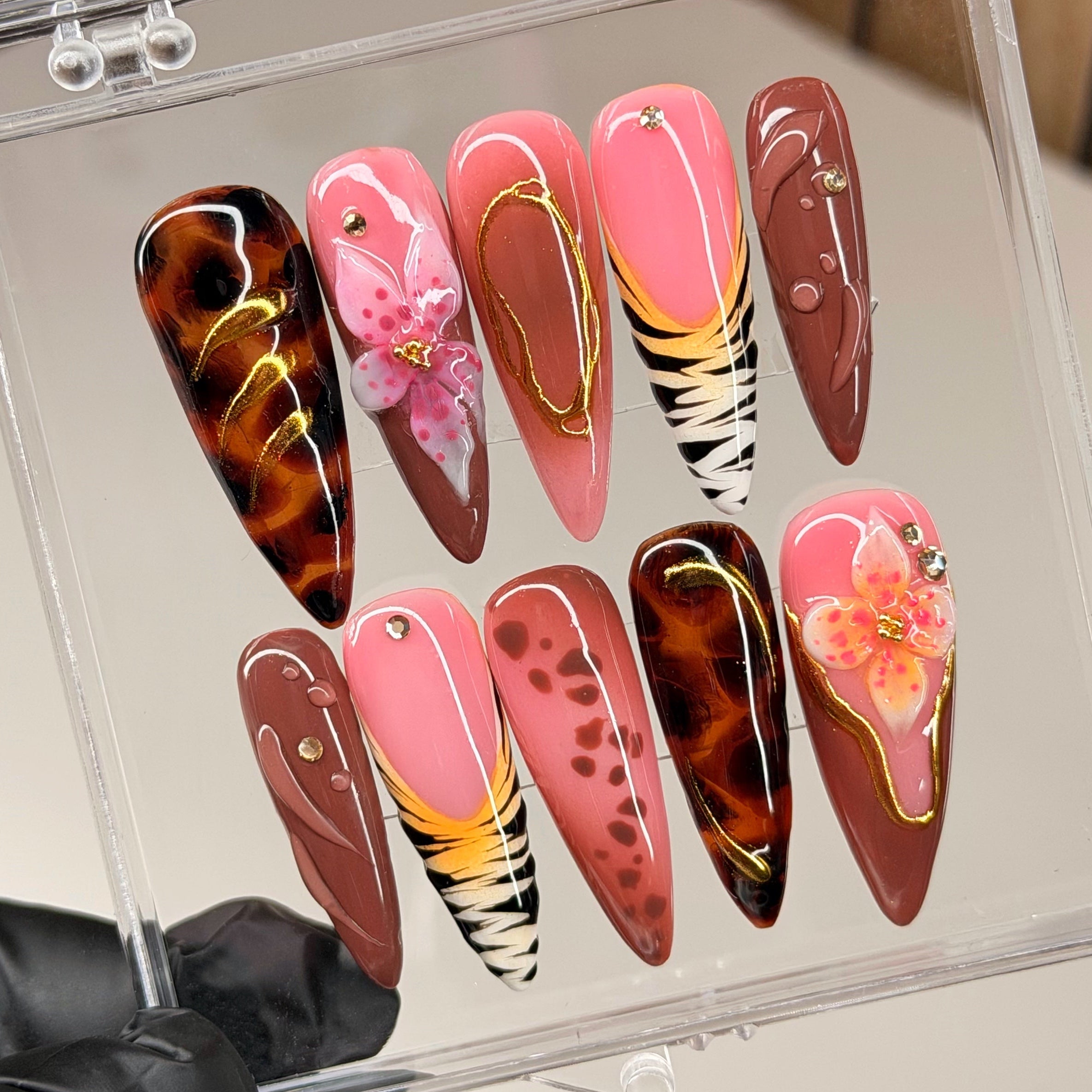 Set of colorful pink, brown, yellow and orange nails. In designs: cheetah print, tortoise shell print, zebra print, flowers, in a clear display case held by a gloved hand.