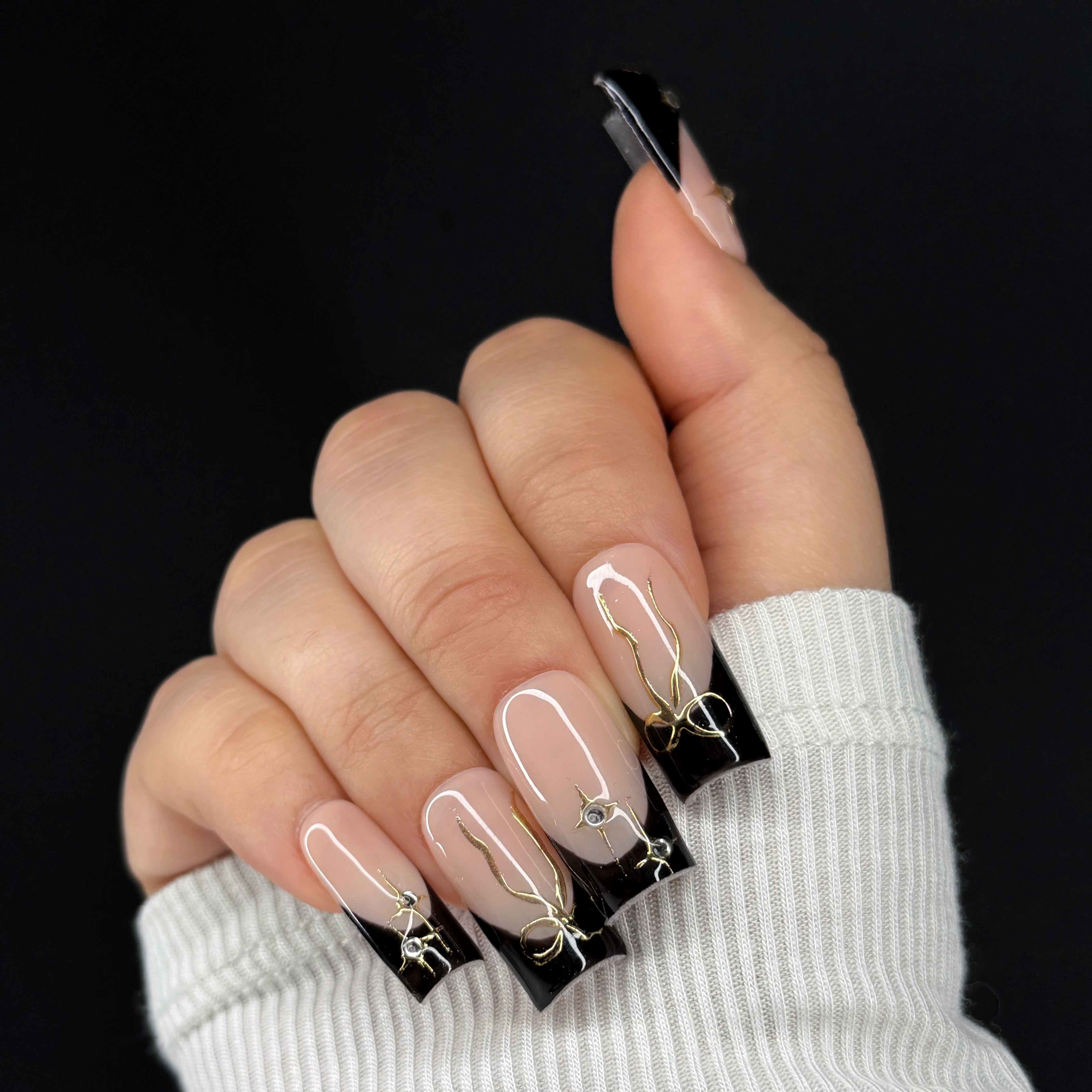 Hand with black French tip nails featuring gold and rhinestone accents on a black background.