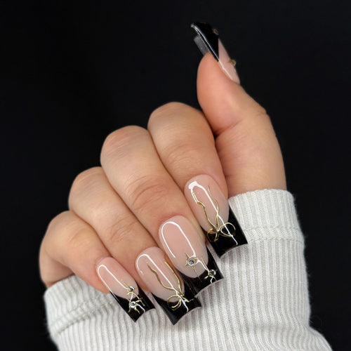 Hand with black French tip nails featuring gold and rhinestone accents on a black background.